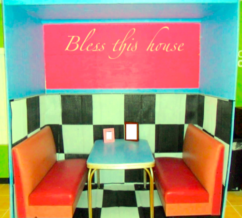 Photograph of a diner booth at Lula Ray's. The booth is bright blue with red seats and with a checkerboard interior. A sign above reads "Bless this house" in script font. Two photographs sit on the table. You cannot see who is in them.
